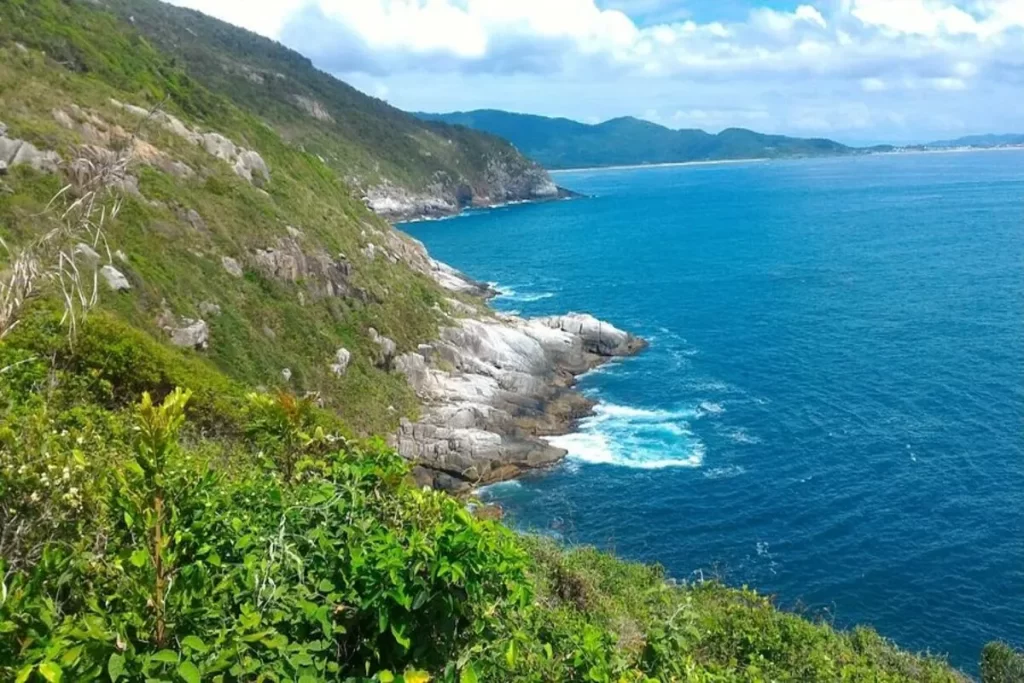 Vista costeira da Praia Lagoinha do Leste, Florianópolis, com morros verdes, falésias rochosas, e mar azul vibrante.