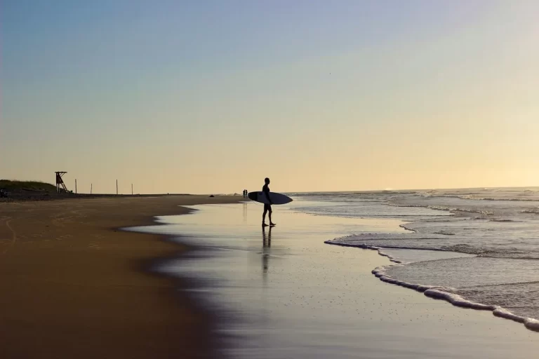 Silhueta de surfista carregando prancha na Praia de Campeche ao entardecer, reflexo na água, ondas e céu dourado.