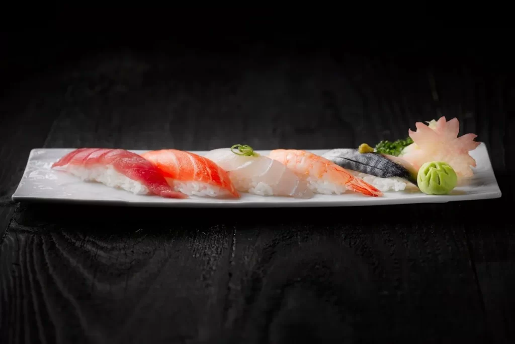 Uma travessa branca com diversos tipos de sushi nigiri, incluindo atum, salmão, peixe branco e camarão, elegantemente dispostos sobre um balcão ou mesa de madeira escura. Acompanham gengibre em conserva e wasabi. restaurante frutos-do-mar