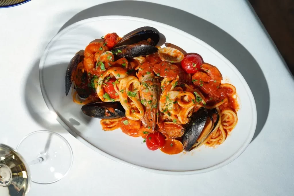 Um prato oval branco com espaguete coberto por um molho vermelho vibrante e uma variedade de frutos do mar, incluindo mexilhões em suas conchas, camarões, lulas e tomate cereja, com um copo de vinho branco parcialmente visível no canto inferior esquerdo.