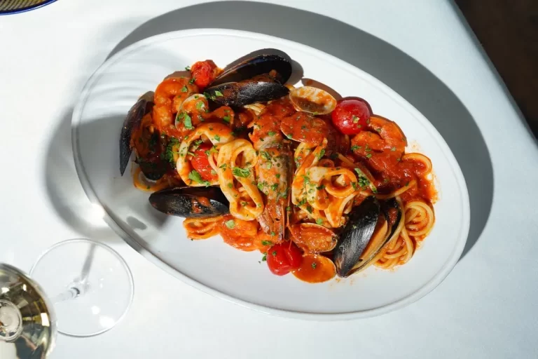 Um prato oval branco com espaguete coberto por um molho vermelho vibrante e uma variedade de frutos do mar, incluindo mexilhões em suas conchas, camarões, lulas e tomate cereja, com um copo de vinho branco parcialmente visível no canto inferior esquerdo.