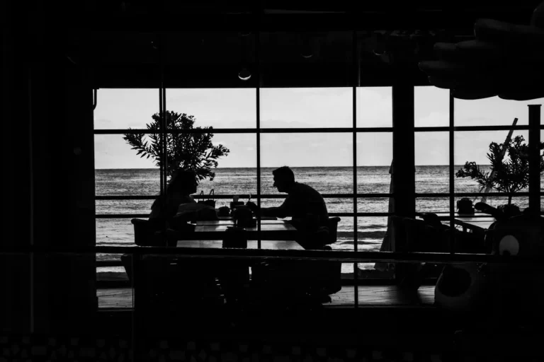 Silhueta de duas pessoas sentadas em um restaurante com grandes janelas, observando o mar em um dia nublado ou ao entardecer, criando uma cena dramática em preto e branco.