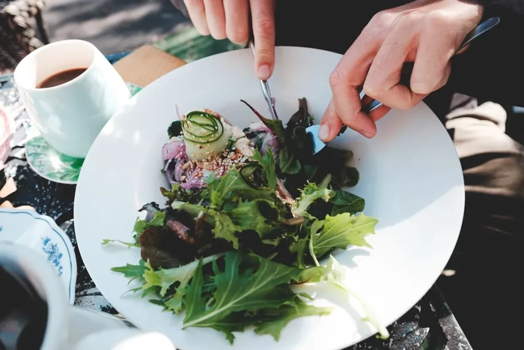 Uma pessoa está usando talheres para comer uma salada fresca e colorida em um prato branco, ao lado de uma xícara de café, representando uma opção saudável e deliciosa de "restaurantes veganos".