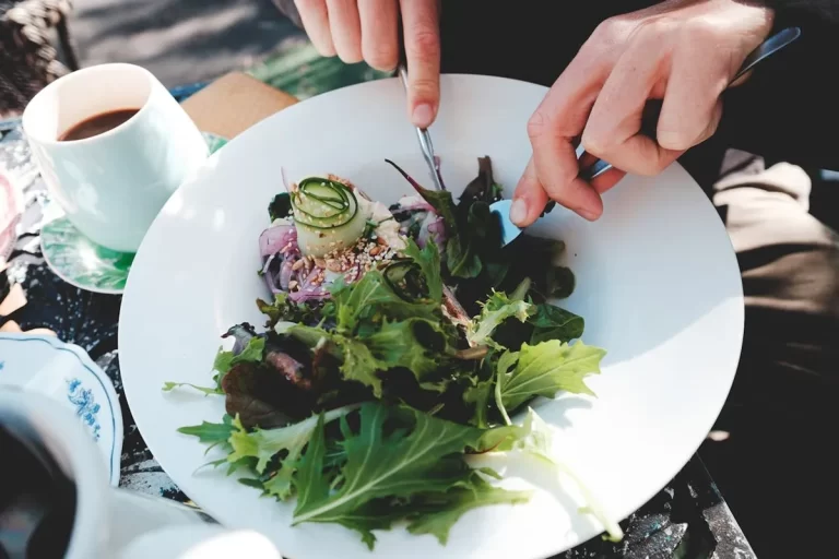 Uma pessoa está usando talheres para comer uma salada fresca e colorida em um prato branco, ao lado de uma xícara de café, representando uma opção saudável e deliciosa de "restaurantes veganos".