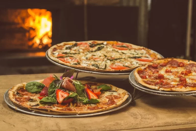 Três pizzas variadas, incluindo uma com morangos, exibidas em um ambiente com forno a lenha, remetendo a um rodízio de pizza em Florianópolis.