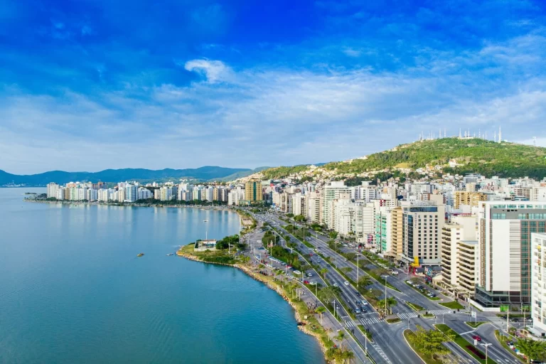 Vista aérea panorâmica da orla de Florianópolis, mostrando a cidade, edifícios à beira-mar, a avenida movimentada e o mar calmo sob um céu azul, refletindo a temperatura em Floripa.
