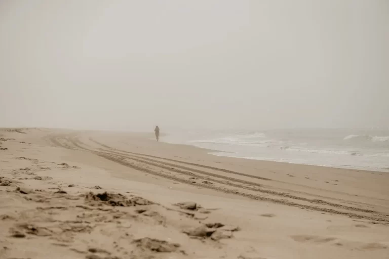 Pessoa caminhando sozinha em uma praia com neblina, com marcas de pneus na areia, na Baía Norte.