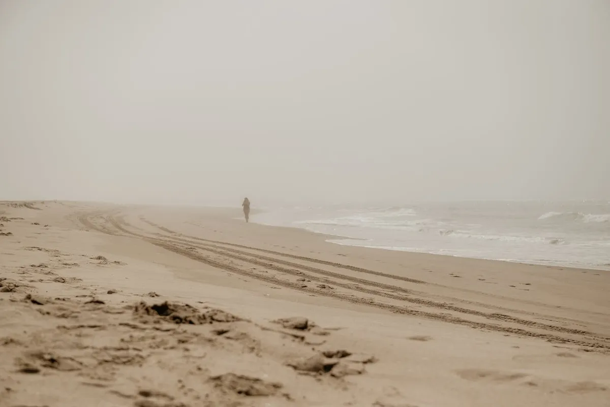Pessoa caminhando sozinha em uma praia com neblina, com marcas de pneus na areia, na Baía Norte.