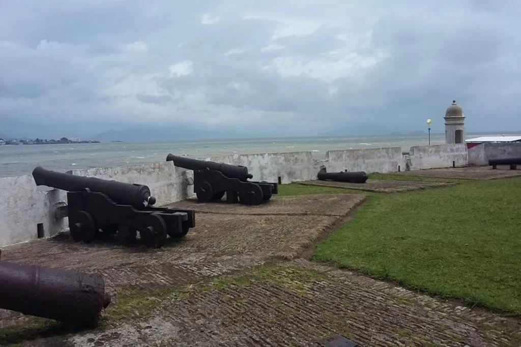 Vista externa do Forte Santana em um dia nublado, com seus canhões antigos alinhados na muralha branca e o mar agitado ao fundo.