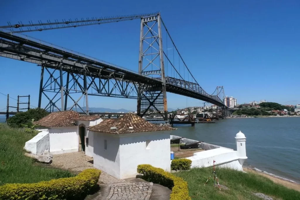 Vista panorâmica do Forte Santana em primeiro plano, com suas pequenas construções brancas de telhado colonial, e a icônica Ponte Hercílio Luz ao fundo, sob um céu azul e límpido.