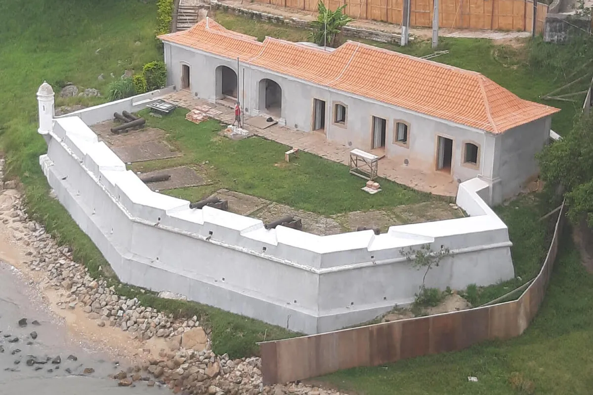 Vista aérea do Forte Santana em Florianópolis, com a estrutura principal e as muralhas brancas em reforma. Canhões antigos estão espalhados pelo gramado verde, e a construção está à beira-mar.