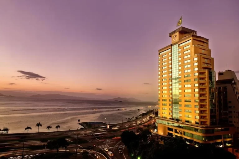 Vista panorâmica do Hotel Majestic Palace Florianópolis durante o crepúsculo. A foto mostra o edifício iluminado com a Beira-Mar Norte, o oceano, a silhueta das montanhas e o céu em tons de roxo e laranja ao entardecer.