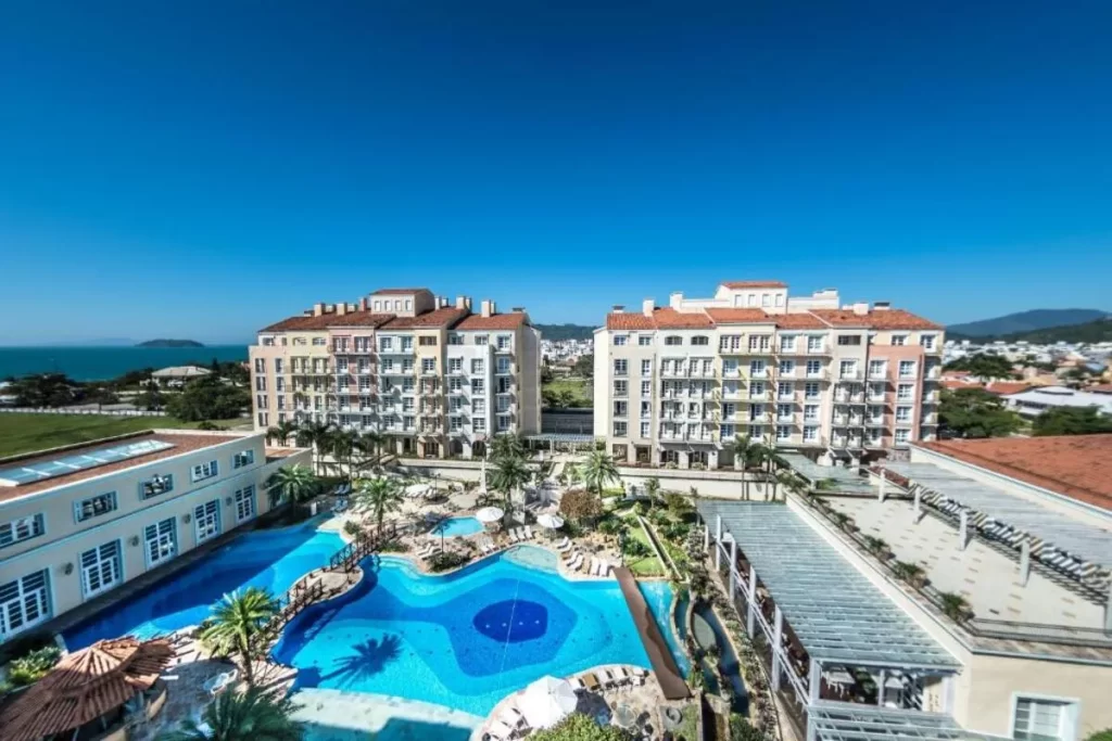 Vista aérea do IL Campanario Villaggio Resort em um dia de sol. A imagem mostra o complexo de piscinas em formato de lagoa, com deck, ponte, palmeiras e a arquitetura em estilo mediterrâneo. Ao fundo, o mar de Jurerê Internacional e a paisagem montanhosa.