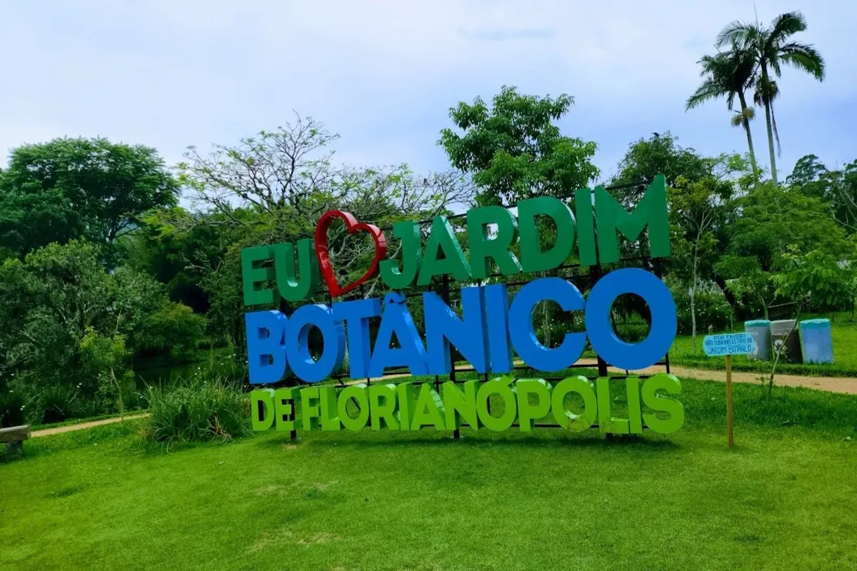 Pessoas caminhando em um dia ensolarado no Jardim Botânico de Florianópolis, por um caminho arborizado com árvores altas e gramado.