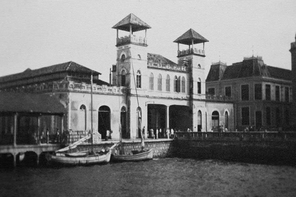 Fotografia em preto e branco do antigo Mercado Público de Florianópolis, com suas torres e arcos. Em primeiro plano, barcos de pesca estão atracados, mostrando a conexão do mercado com o mar e a cultura local da época.