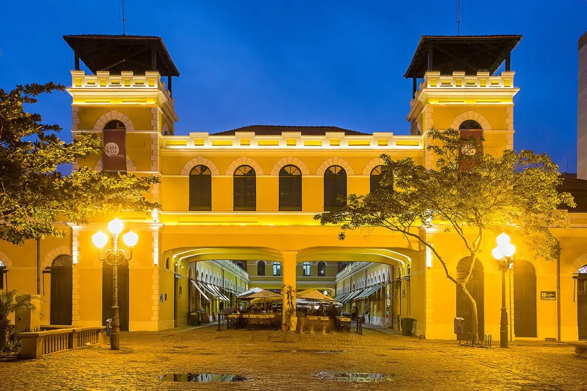 Fachada noturna do Mercado Público de Florianópolis, iluminada e em destaque sob um céu azul-escuro. A imagem mostra as duas torres do prédio e a passagem que leva ao pátio interno, com suas luzes e mesas de restaurantes.