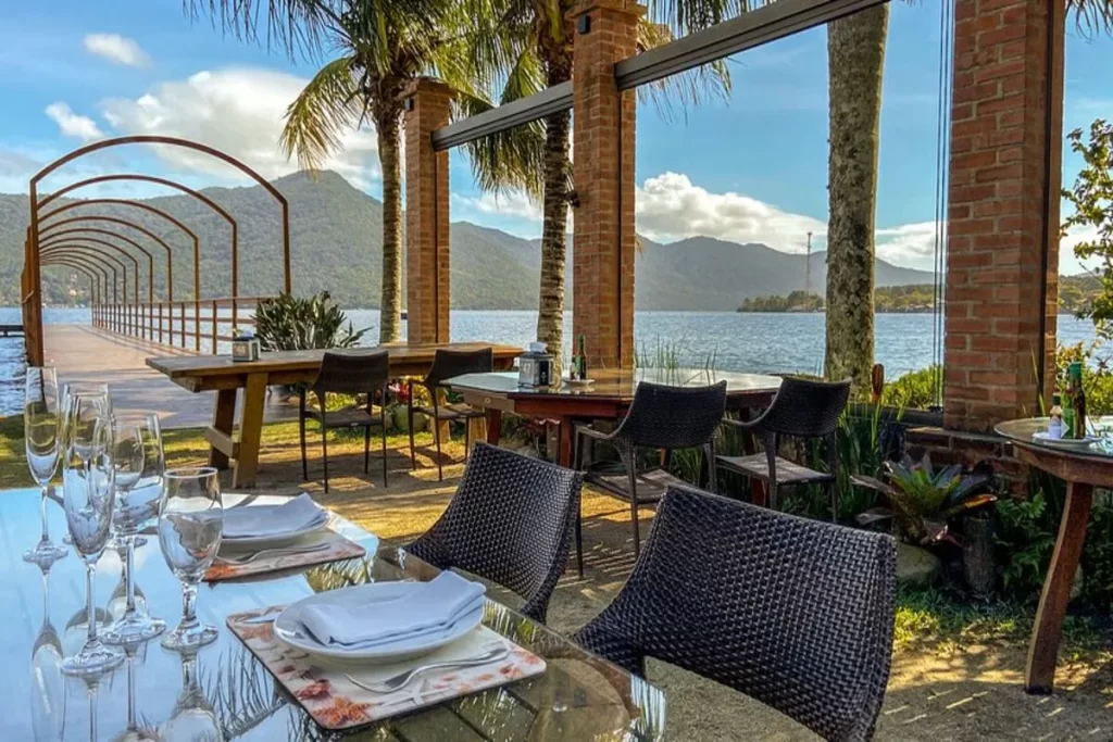 Vista de mesas de restaurante e um píer de madeira à beira de um lago, com montanhas ao fundo, no local conhecido como Ponta das Caranhas.