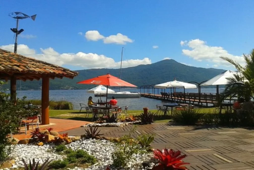 Vista panorâmica do Restaurante Ponta das Caranhas, com mesas ao ar livre, píer sobre a água e montanha ao fundo, sob um céu azul.