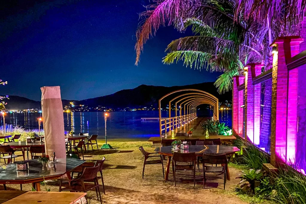 Vista noturna do restaurante Ponta das Caranhas, com píer iluminado, mesas à beira d'água e luzes coloridas, criando uma atmosfera romântica.
