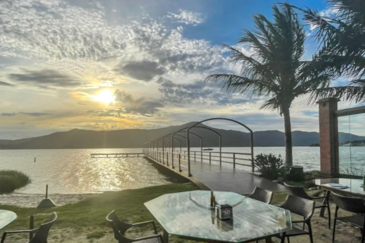 Vista panorâmica do Restaurante Ponta das Caranhas ao pôr do sol, com mesas à beira de um lago, píer de madeira e montanhas ao fundo.