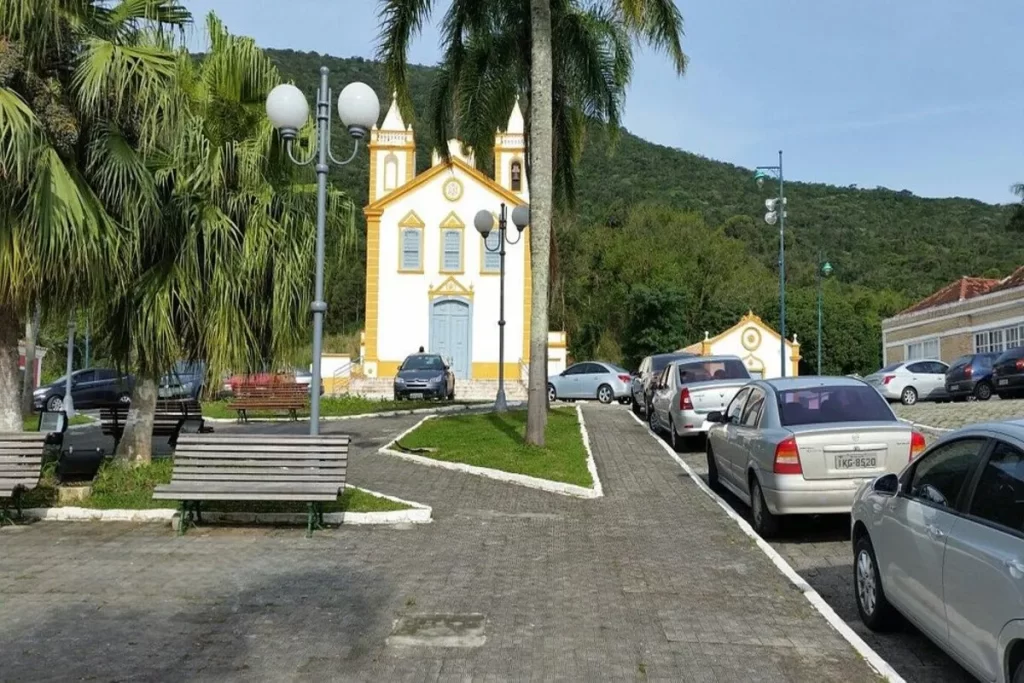Vista da praça principal do Ribeirão da Ilha, com a Igreja de Nossa Senhora da Lapa e a arquitetura açoriana, em um dia ensolarado.