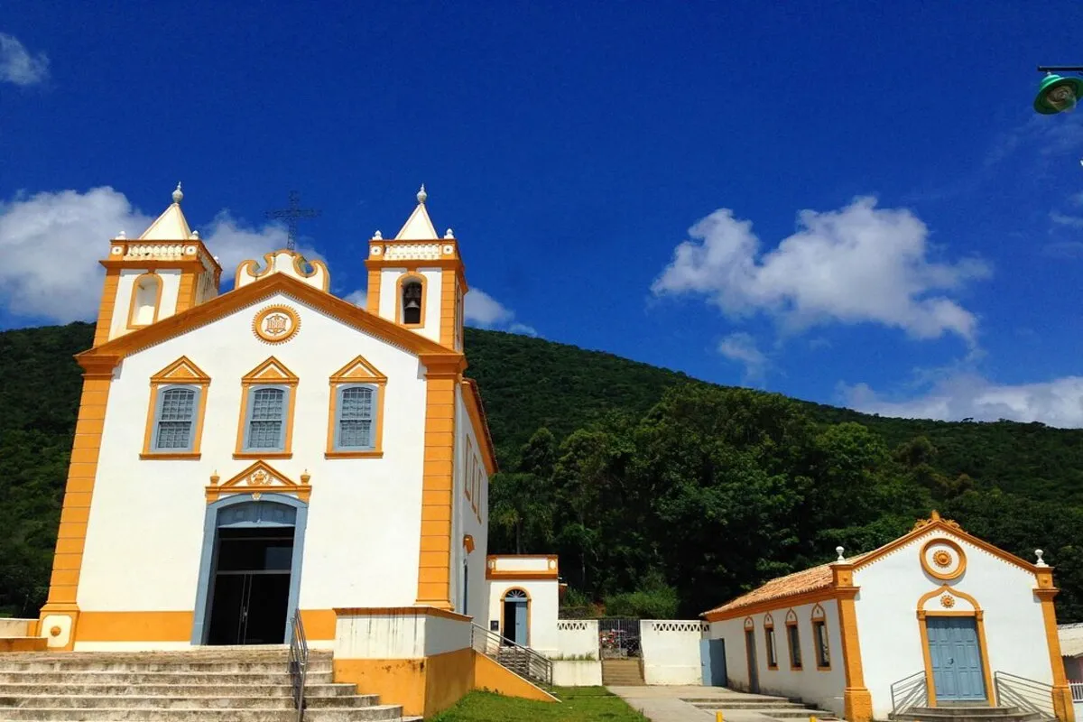 A Igreja de Nossa Senhora da Lapa no Ribeirão da Ilha, com suas fachadas brancas e detalhes em laranja, e a vegetação exuberante ao fundo sob um céu azul e com nuvens.