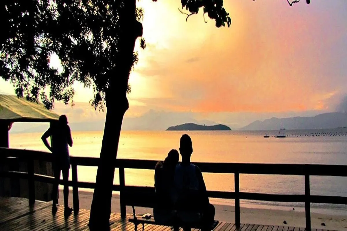 Silhueta de casal em deck assistindo ao pôr do sol sobre a baía de Sambaqui, em Florianópolis.