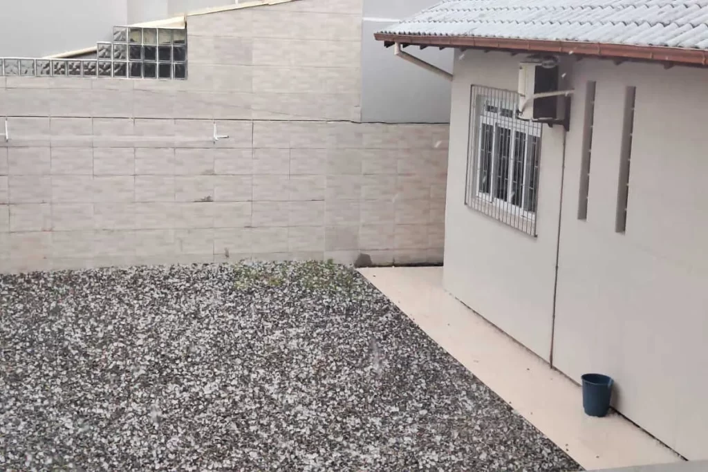 Cena de uma intensa chuva de granizo caindo em um quintal, com o chão coberto por uma espessa camada de gelo.