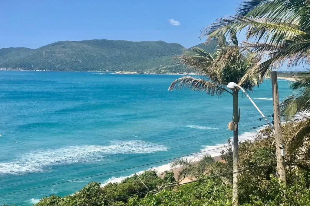 Vista panorâmica da praia, com o mar azul-turquesa, areia clara e a vegetação tropical. Ao fundo, uma montanha com densa vegetação e a praia da Armação. A imagem representa a vista do Mirante Morro das Pedras.