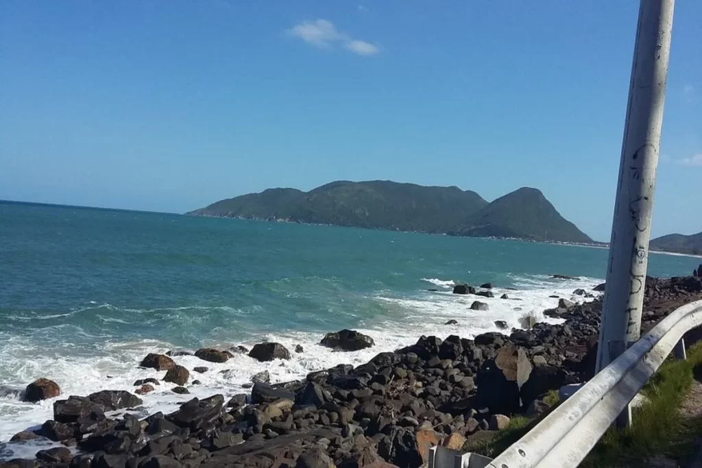 Vista panorâmica do Mirante Morro das Pedras, com o mar agitado quebrando nas pedras, a Ilha do Campeche ao fundo e a estrada com um guarda-corpo no primeiro plano.