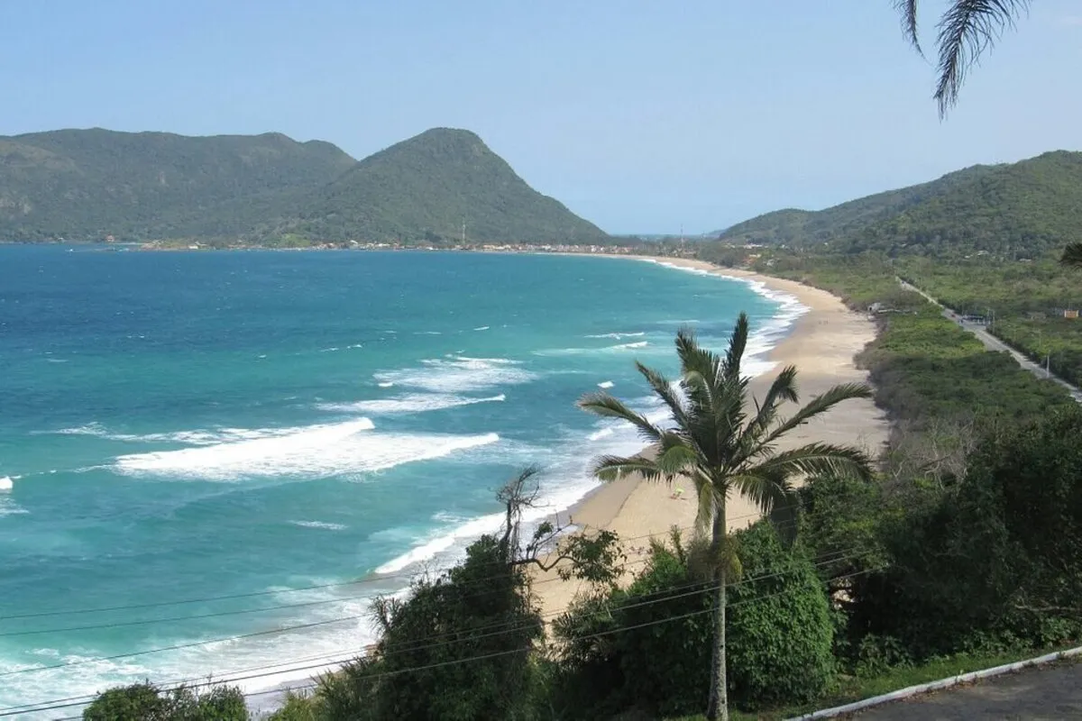 Vista panorâmica da praia, com o mar azul-turquesa, areia clara e a vegetação tropical. Ao fundo, uma montanha com densa vegetação e a praia da Armação. A imagem representa a vista do Mirante Morro das Pedras.