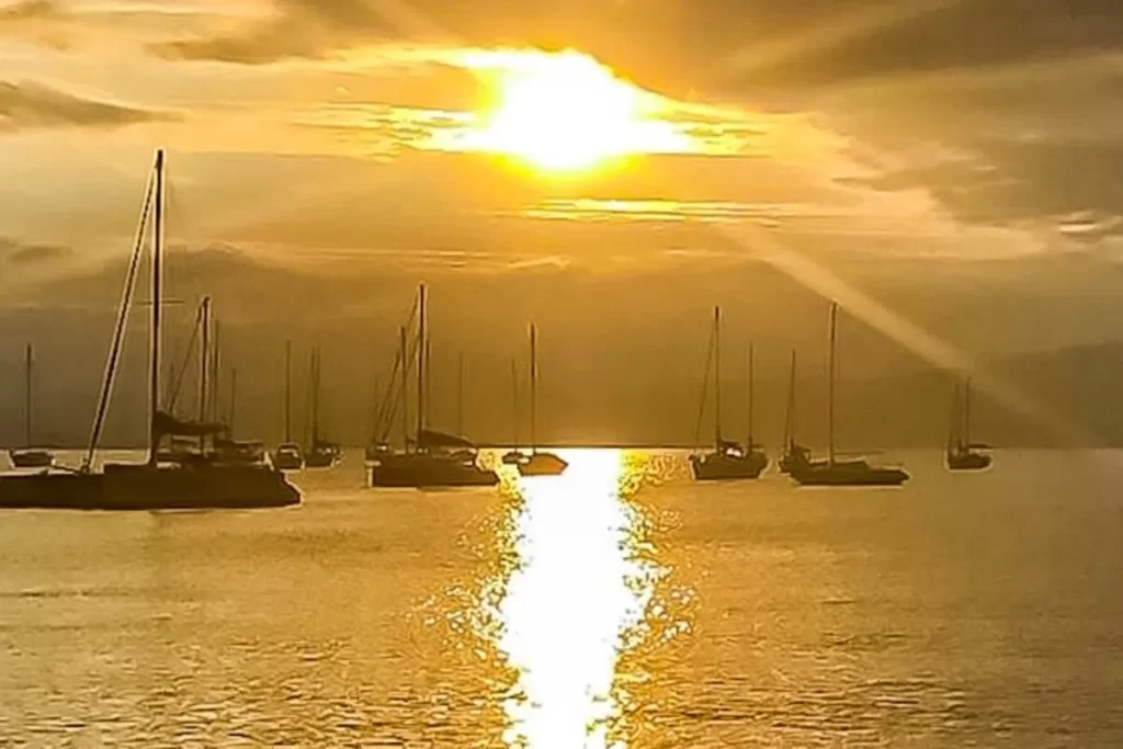 Um pôr do sol em Santo Antônio de Lisboa, onde o céu e o mar se tingem de dourado. Diversos veleiros e barcos estão ancorados, criando silhuetas contra a luz intensa do sol.