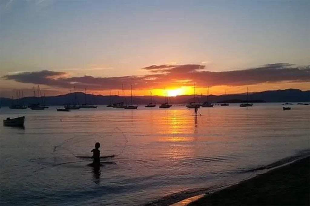Silhueta de um pescador na água arremessando uma rede, com vários barcos de pesca e veleiros ancorados ao fundo, durante um belo pôr do sol em Santo Antônio de Lisboa, Florianópolis.