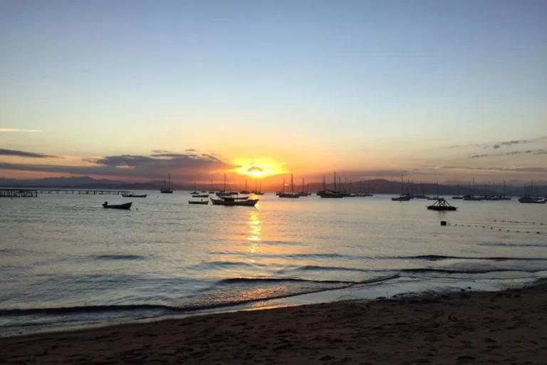 Um pôr do sol em Santo Antônio de Lisboa com barcos ancorados na água calma. A praia de areia e o reflexo do sol no mar compõem a cena tranquila.