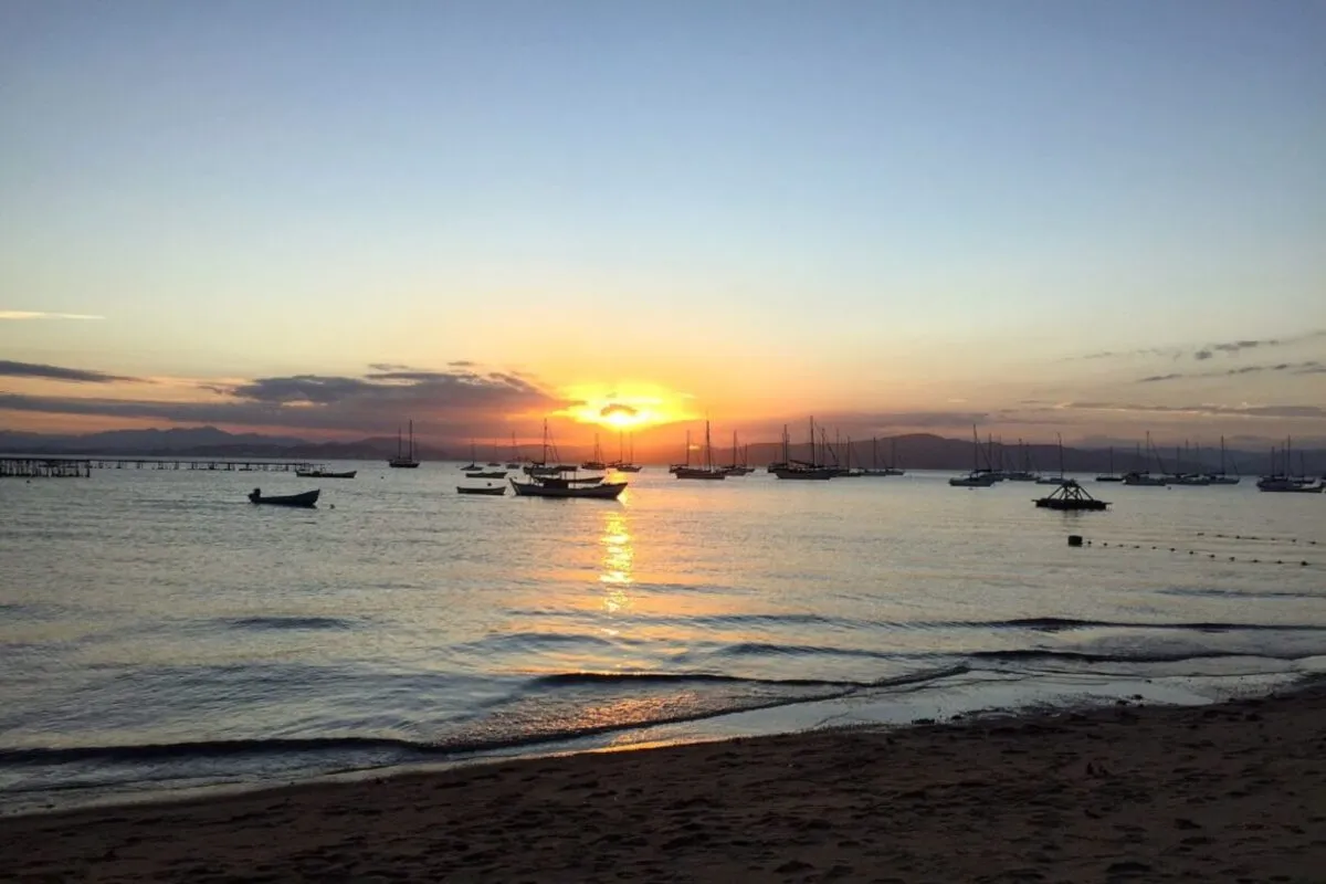 Um pôr do sol em Santo Antônio de Lisboa com barcos ancorados na água calma. A praia de areia e o reflexo do sol no mar compõem a cena tranquila.