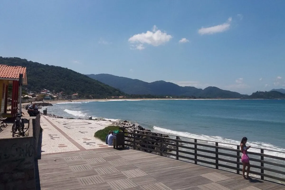Vista panorâmica do calçadão e trapiche da Praia da Armação, com o mar, montanhas e casas da vila ao fundo.