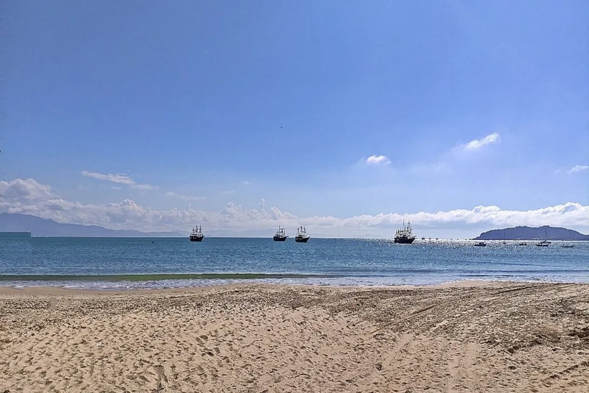 A praia da Cachoeira do Bom Jesus, em Florianópolis, com areia clara, mar calmo e barcos ao longe, sob um céu azul.