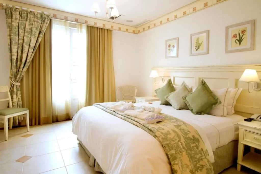 Interior de um quarto de resort all inclusive em Florianópolis, com cama king-size, móveis de madeira pintada de branco, roupa de cama em tons claros com detalhes florais em verde, cortinas em tons de bege e verde, e piso de cerâmica. A imagem transmite um ambiente clássico e romântico. resorts all inclusive Florianópolis