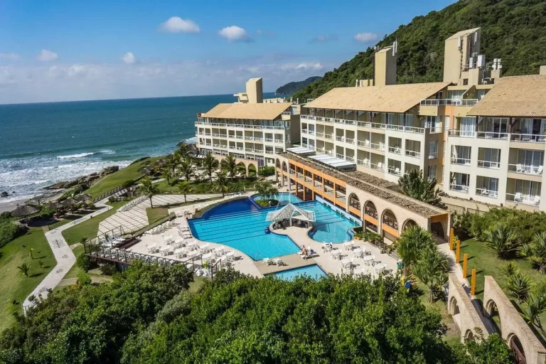 Vista aérea de um resort all inclusive em Florianópolis. A foto mostra o edifício do hotel com telhados de cerâmica, piscina com borda infinita, palmeiras e espreguiçadeiras. Ao fundo, o mar com ondas, a costa rochosa e a vegetação nativa. resorts all inclusive Florianópolis