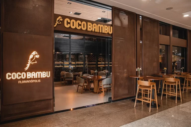 A entrada do restaurante Coco Bambu em Florianópolis, mostrando a fachada de vidro, o logo na parede de metal escuro, os painéis laterais perfurados e as mesas de madeira na área externa.