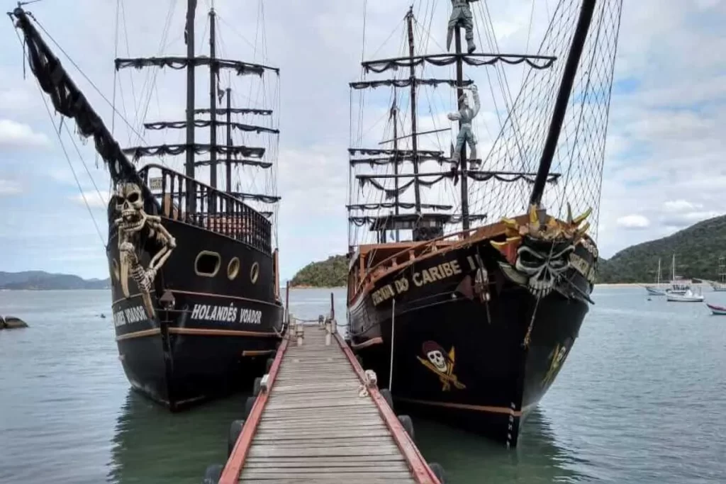 Duas escunas temáticas de barco pirata, "Holandês Voador" e "Escuna do Caribe", atracadas lado a lado em um pier de madeira em águas tranquilas. Ambas têm caveiras decorativas na proa e mastros imponentes, sob um céu nublado.
