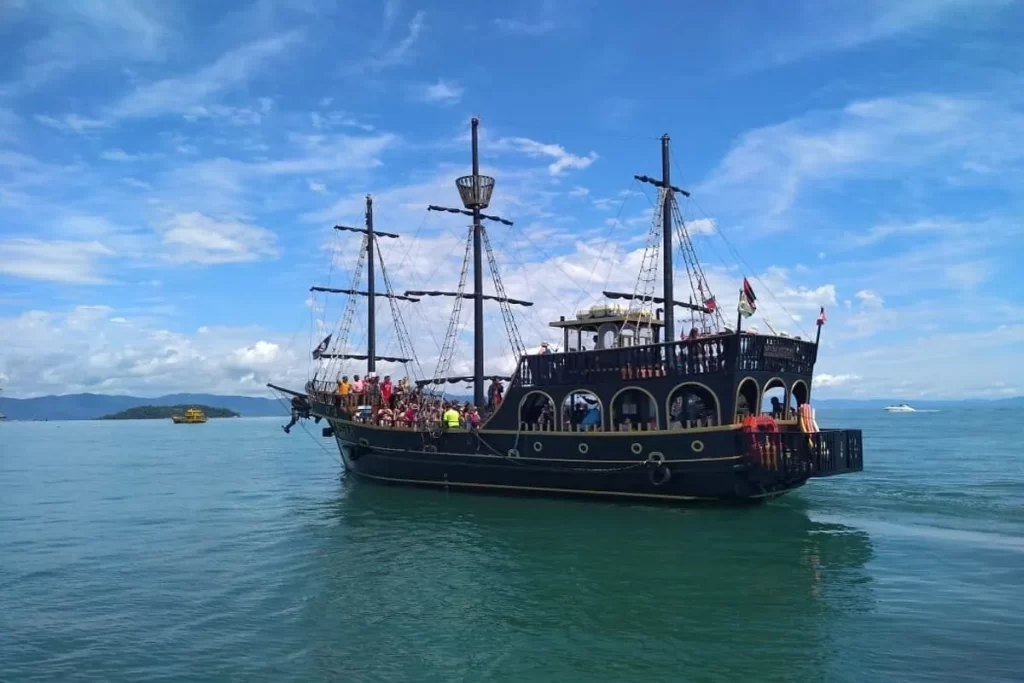 Vista lateral de uma grande escuna de cor escura, semelhante a um barco pirata, navegando em águas calmas. A embarcação está cheia de turistas no convés sob um céu azul com nuvens brancas e esparsas. Ao fundo, um morro verde e outras embarcações podem ser vistas.