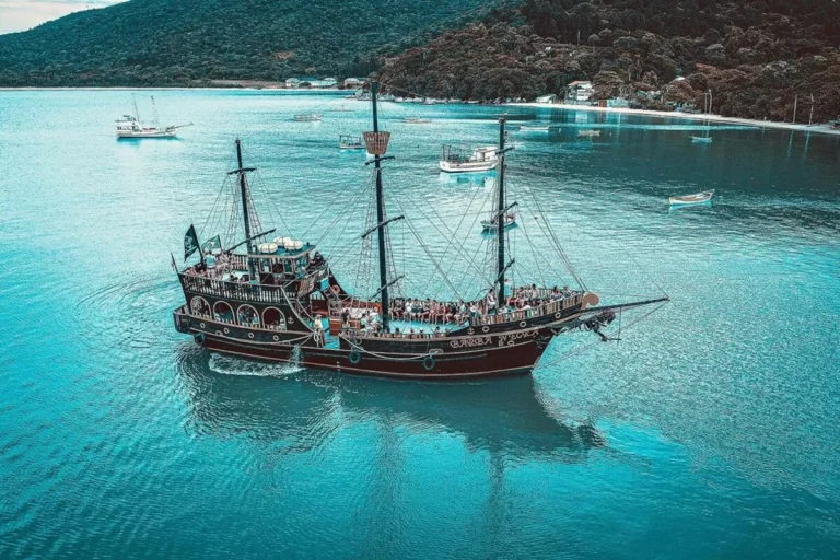 Vista aérea de uma grande escuna temática de barco pirata navegando em águas azuis claras e calmas. A embarcação, cheia de turistas, está em frente a uma costa com um morro coberto por mata atlântica densa, casas e outras pequenas embarcações.