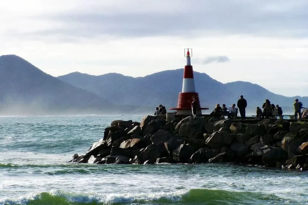 Farol da Barra da Lagoa em Florianópolis, com pessoas observando o mar e montanhas ao fundo