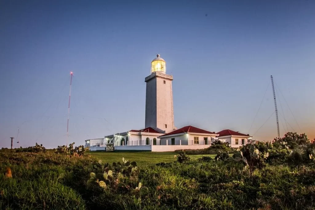 Farol de Santa Marta, uma torre branca com luz acesa no topo, ao anoitecer. O farol está em uma colina gramada, com cactos no primeiro plano e o céu em tons de azul e laranja. A estrutura é ladeada por edifícios brancos com telhados vermelhos.Uma foto do Farol de Santa Marta no momento do crepúsculo. O céu azul escuro no topo da imagem se mistura com tons alaranjados e amarelados perto do horizonte. O farol, uma torre branca e cilíndrica, está iluminado e com sua luz acesa, destacando-se contra a escuridão crescente. Na base do farol, há edifícios brancos com telhados de telha vermelha. O farol está localizado em um terreno com grama verde e cactos espalhados em primeiro plano. A imagem transmite uma sensação de tranquilidade e segurança, com a luz do farol brilhando na paisagem de transição entre o dia e a noite.