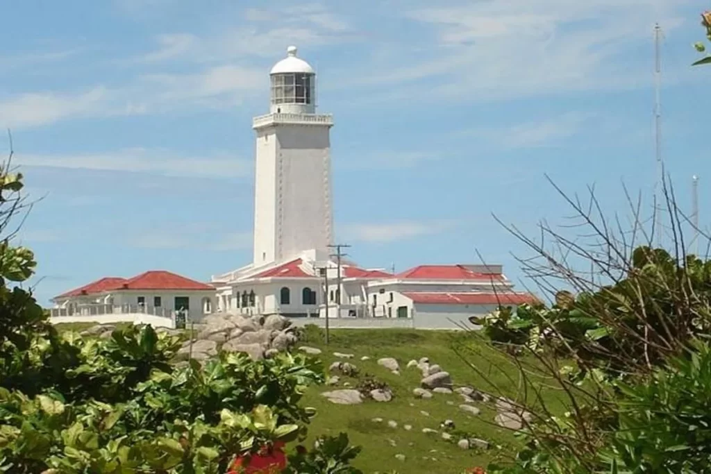 Vista do Farol de Santa Marta, uma estrutura cilíndrica branca, com sua cúpula de vidro no topo, sobre uma colina rochosa. Edifícios brancos com telhados de telha vermelha estão na base do farol. O céu é azul com nuvens, e a paisagem é verdejante com pedras grandes e plantas no primeiro plano.