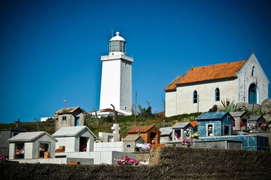 Vista do Farol de Santa Marta, uma torre branca no topo de uma colina, com um cemitério em primeiro plano. O cemitério possui vários túmulos coloridos e uma igreja de fachada branca e telhado vermelho no fundo. O céu é de um azul intenso e o sol é forte.