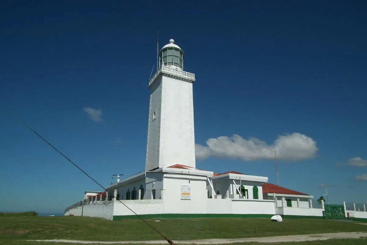 Farol de Santa Marta, uma torre branca com cúpula cinza-clara, em um dia ensolarado. A estrutura está sobre uma colina gramada, com prédios brancos de telhado vermelho na base. O céu é azul intenso com poucas nuvens brancas, e o farol é visto em uma perspectiva levemente inclinada.