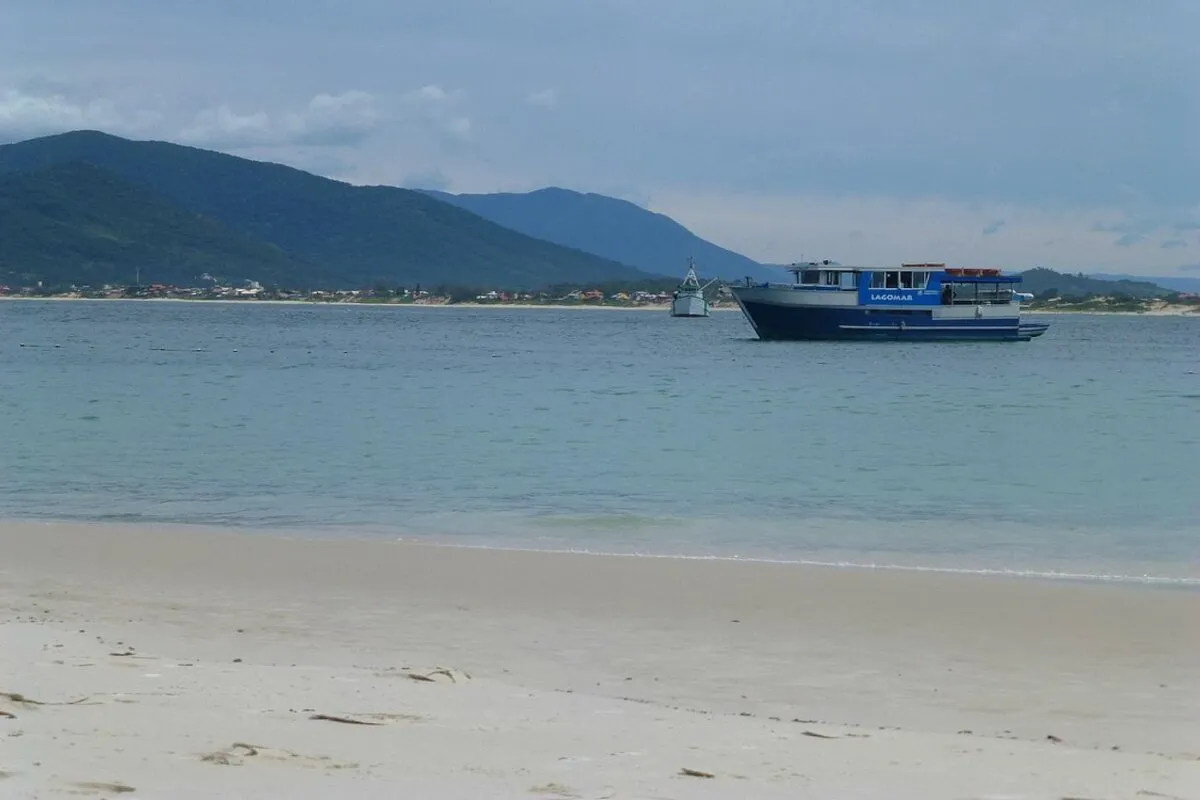 Barco de transporte turístico ancorado próximo à Ilha do Campeche, em Florianópolis, com vista para o mar e montanhas ao fundo.