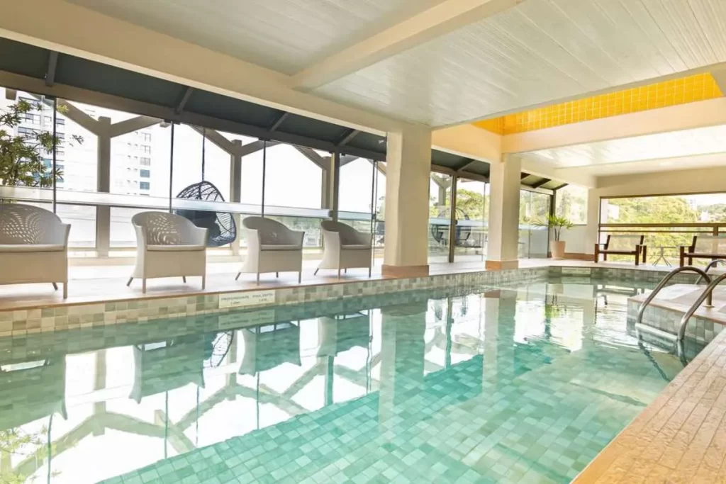 A área da piscina interna do hotel Mercure Florianópolis, mostrando a piscina com azulejos claros, cadeiras brancas de vime, cadeiras de balanço suspensas e grandes janelas de vidro com vista.